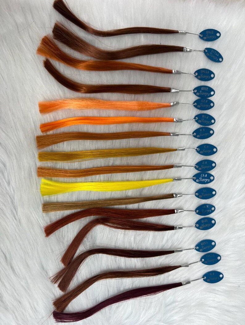 May include: A collection of hair swatches in various shades of brown, orange, and yellow. Each swatch is attached to a small metal loop with a blue tag. The tags have white text. The swatches are arranged on a white, textured surface.