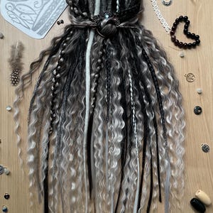May include: A collection of black, grey, and white dreadlocks, adorned with a decorative hair accessory. The dreads are displayed on a wooden surface, accompanied by feathers and beads. The style evokes a bohemian aesthetic.