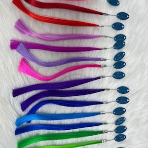 May include: A collection of colorful hair extensions in shades of pink, red, purple, blue, and green. Each extension is attached to a small metal clip with a blue tag. The extensions are arranged on a white, textured surface.