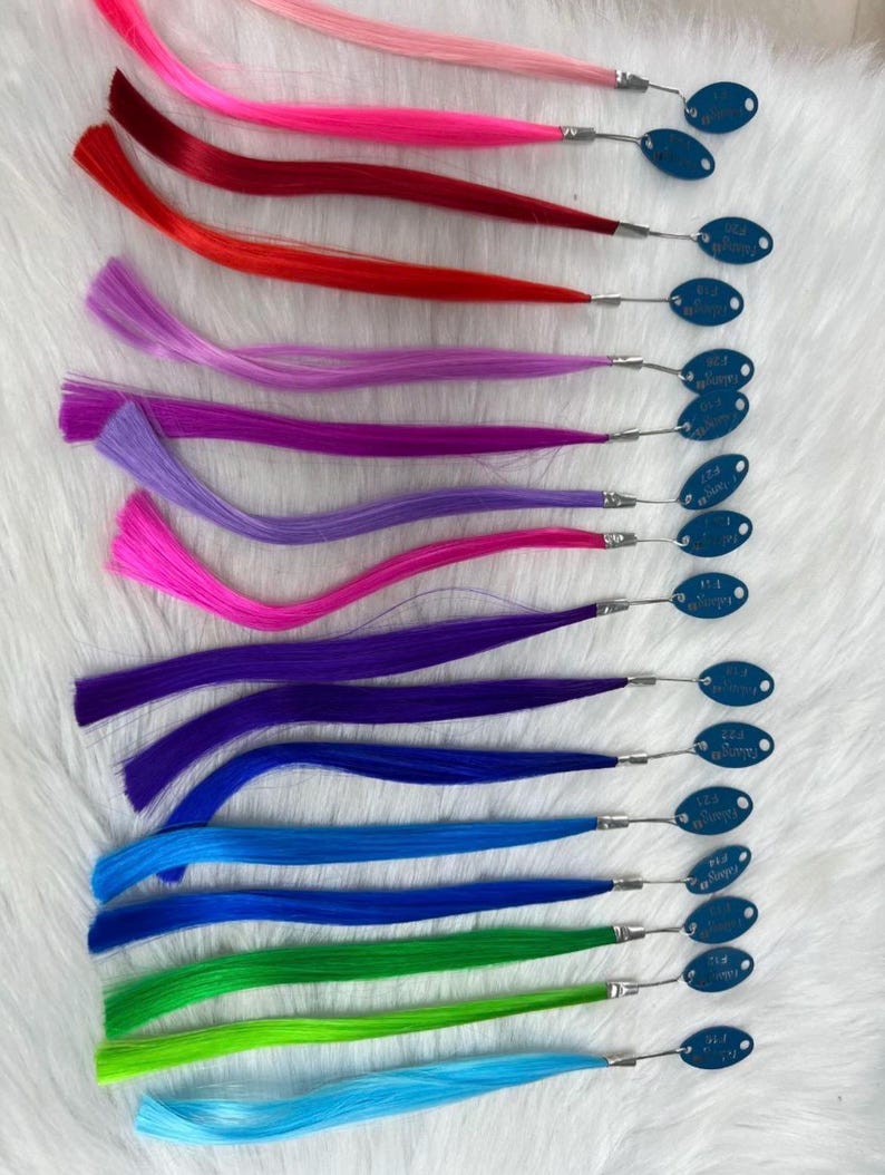 May include: A collection of colorful hair extensions in shades of pink, red, purple, blue, and green. Each extension is attached to a small metal clip with a blue tag. The extensions are arranged on a white, textured surface.