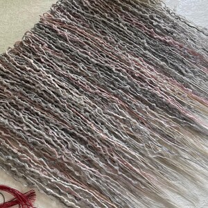 May include: A set of synthetic hair extensions featuring a blend of grey, white, and soft pink tones with a textured, wavy finish. The strands are arranged in parallel rows, accompanied by a small red pendant on a cord and a separate red tassel.