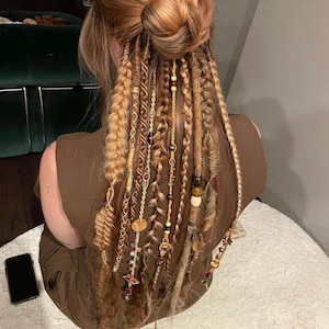 May include: A person with long brown hair with multiple strands of blonde and brown synthetic dreadlocks with beads and charms. The dreadlocks are pulled back into a bun.