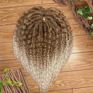 May include: A long, curly hair extension with a light brown to white ombre color. The hair is tied together with a brown string and a small, round, brown and white stone charm.