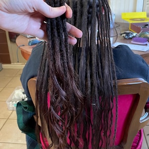 May include: Close-up of dark brown dreadlocks with reddish-brown and pink highlights. The dreadlocks are being held up, showcasing their length and texture. The hair is styled with metal clips and is partially installed.