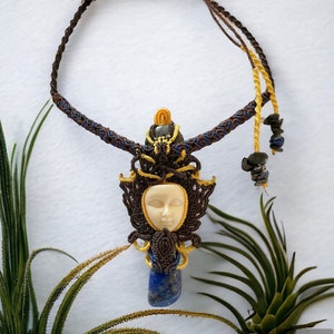 May include: A macrame necklace with a carved face pendant and a blue lapis lazuli stone. The necklace is made with brown, blue, and yellow cords.