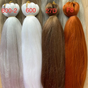 May include: A selection of synthetic braiding hair bundles in shades of blonde, grey, white, brown, and orange. Each bundle is labelled with a number and colour code, such as "16S" and "27D". The hair is secured at the top with a yellow elastic band.