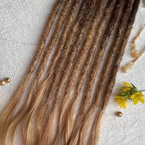 May include: A set of synthetic dreadlocks featuring a color gradient that transitions from dark brown at the top to a light blonde at the ends. The dreadlocks have a textured, matted appearance with loose, straight hair strands at the bottom tips.