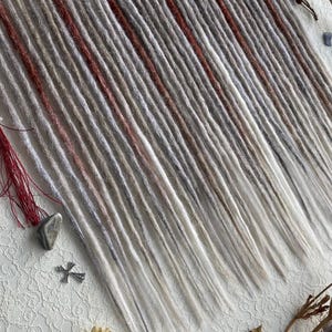 Pode incluir: Uma coleção de dreadlocks feitos à mão em tons de cinza, branco e marrom avermelhado. Os dreads estão dispostos sobre uma superfície de renda branca, com elementos decorativos como flores secas e pequenos amuletos.