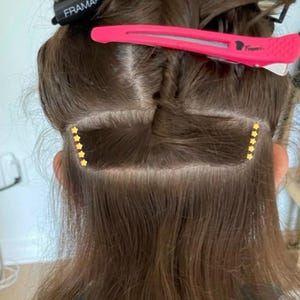 May include: A person's brown hair with two black hair clips, one with the text "FRAMAR" and the other with the text "Taper", and two rows of gold star-shaped hair extensions.