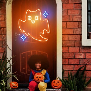 May include: A young person dressed in a Halloween costume holds a carved pumpkin while sitting on the steps of a brick house decorated with a glowing neon ghost.