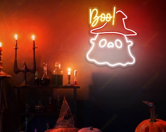Op de afbeelding: Een wit neonbord met een geest die een heksenhoed draagt en het woord "Boo!" in geel neon. Het bord hangt aan een rode muur met kaarsen en pompoenen op de voorgrond.