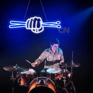 Op de afbeelding: Een blauw neonbord in de vorm van een vuist die drumstokken vasthoudt, hangt boven een drummer die een drumstel bespeelt. Het bord is wit met een blauwe omtrek. De drummer draagt een beige overhemd.
