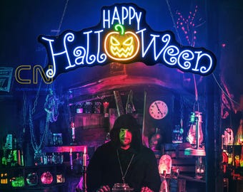 Op de afbeelding: Een neonbord met de woorden "Happy Halloween" in blauw en groen neonlicht. Het bord heeft een gestileerde pompoen met een griezelig gezicht in het midden. Het bord hangt aan een ketting en wordt verlicht tegen een donkere achtergrond.