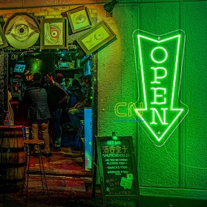 May include: A neon green "OPEN" sign in the shape of an arrow is lit up on a green wall. The entrance to a bar is visible, with people inside. A chalkboard sign lists "ART BAR" and other Japanese text.