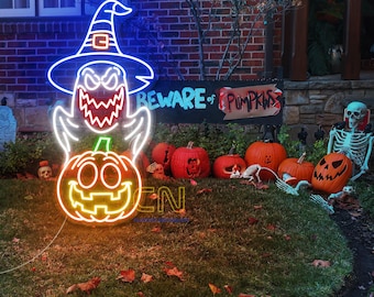 Op de afbeelding: Een neon Halloween-decoratie met een spook met een heksenhoed en een pompoen. Het bord luidt "BEWARE OF PUMPKINS" en is omgeven door gesneden pompoenen en een skelet. Het woord "OPEN" is ook zichtbaar.