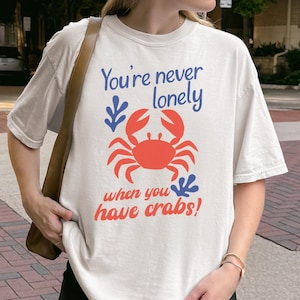May include: An off-white short-sleeve t-shirt featuring a graphic of a red crab centered on the chest. The text reads "You're never lonely when you have crabs!" in blue and red lettering, accompanied by two small blue leaf-like illustrations.