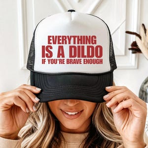 Op de afbeelding: Een wit en zwart truckerpetje met een schuimrubberen voorpaneel waarop de rode tekst "EVERYTHING IS A DILDO IF YOU'RE BRAVE ENOUGH" in een vet, schreefloos lettertype is gedrukt. De pet heeft een zwarte klep en een zwart mesh achterpaneel.