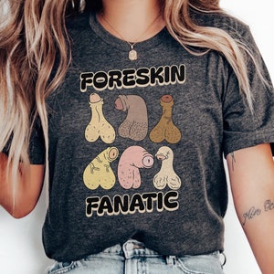 May include: A dark grey heather t-shirt featuring the text "FORESKIN FANATIC" in a bold, retro-style font. The graphic includes six stylized, cartoon-like illustrations of penises in various skin tones arranged in two rows on the chest.