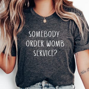 May include: A dark grey t-shirt with the text "Somebody order womb service?" printed in white.