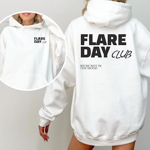 May include: A white hoodie with a front pocket and a drawstring hood. The front of the hoodie has the text "FLARE DAY CLUB" and "WE'RE NOT IN THE MOOD" printed in black. The back of the hoodie has the same text.