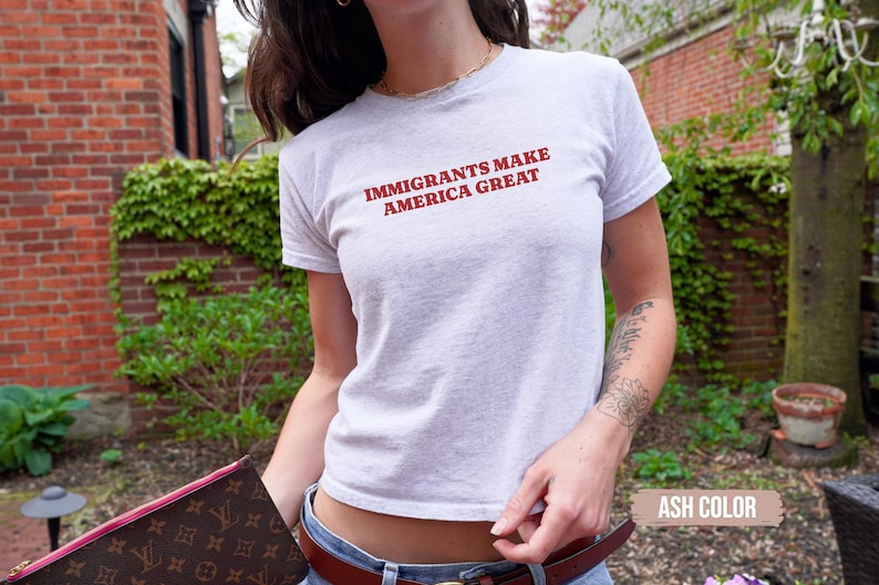 May include: Ash-colored, short-sleeved t-shirt with the red text "IMMIGRANTS MAKE AMERICA GREAT." The shirt is worn by a person. The person is holding a brown and pink patterned clutch. The background includes a brick wall and greenery.
