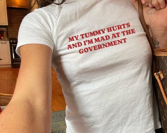 May include: White t-shirt with the red text "MY TUMMY HURTS AND I'M MAD AT THE GOVERNMENT." The shirt is a short-sleeved, fitted style. The person is wearing blue jeans and carrying a brown handbag.