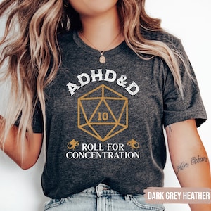 May include: A dark grey heather t-shirt with the text "ADHD&D Roll for Concentration" and a gold graphic of a twenty-sided die with the number 10 on it.