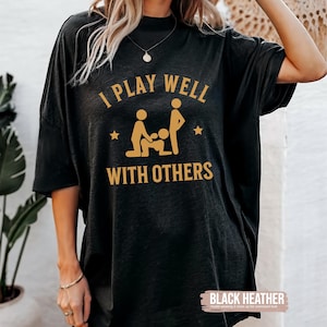 Könnte beinhalten: Schwarzes Heather-Oversize-T-Shirt mit dem goldenen Text "I PLAY WELL WITH OTHERS". Die Grafik zeigt Strichmännchen. Das Shirt ist ein lässiger Unisex-Stil.