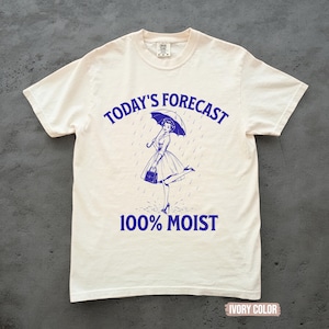 May include: An ivory short-sleeve t-shirt featuring a blue graphic print. The design shows a person holding an umbrella in the rain with the text "TODAY'S FORECAST" arched above and "100% MOIST" printed below the illustration in a bold blue font.