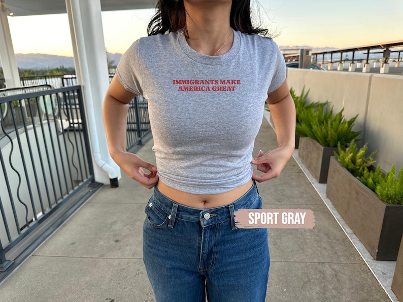 May include: A light gray t-shirt with the text "IMMIGRANTS MAKE AMERICA GREAT" in red. The shirt is paired with blue jeans. The words "SPORT GRAY" are in a rectangular box in the lower right corner of the image.
