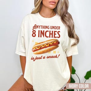 May include: Ivory-colored t-shirt with a hot dog graphic and the text "Anything under 8 inches is just a snack!" The hot dog has mustard and is in a bun. The shirt is a casual style.