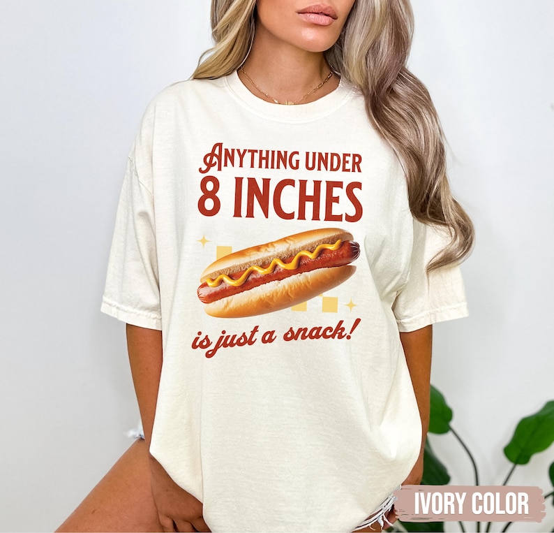 May include: Ivory-colored t-shirt with a hot dog graphic and the text "Anything under 8 inches is just a snack!" The hot dog has mustard and is in a bun. The shirt is a casual style.