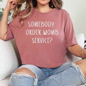 Op de afbeelding: Een vrouw draagt een mauve t-shirt met de tekst "Somebody order womb service?" in witte letters. Ze zit op een witte bank met een zwart-wit geblokte deken.