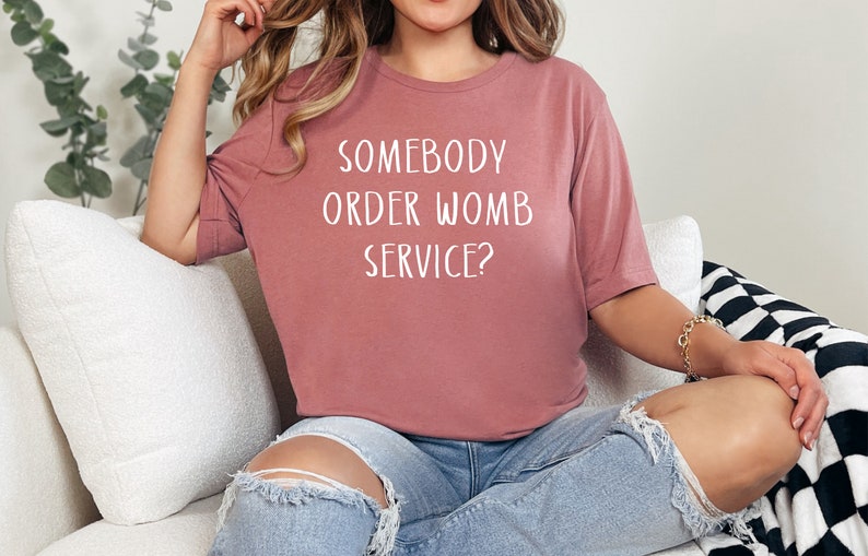 Op de afbeelding: Een vrouw draagt een mauve t-shirt met de tekst "Somebody order womb service?" in witte letters. Ze zit op een witte bank met een zwart-wit geblokte deken.
