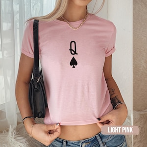 Może przedstawiać: Jasnoróżowa koszulka z czarną grafiką Królowej Pik. Koszulka ma krótkie rękawy i okrągły dekolt. Tekst "LIGHT PINK" jest widoczny na dole zdjęcia.