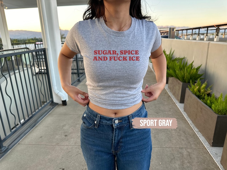 May include: A grey t-shirt with the text "SUGAR, SPICE AND FUCK ICE" in red. The shirt is worn with blue jeans. The words "SPORT GRAY" are in a white box.