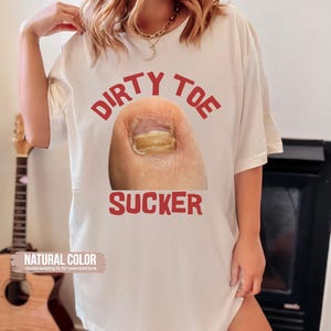 Pu&ograve; includere: T-shirt color crema con le parole "DIRTY TOE SUCKER" in lettere rosse, sopra e sotto un'immagine ravvicinata di un'unghia del piede. La maglietta &egrave; etichettata "NATURAL COLOR" ed &egrave; indossata per un look oversize.