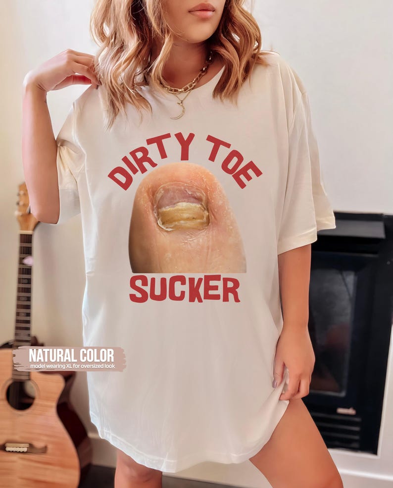 Pu&ograve; includere: T-shirt color crema con le parole "DIRTY TOE SUCKER" in lettere rosse, sopra e sotto un'immagine ravvicinata di un'unghia del piede. La maglietta &egrave; etichettata "NATURAL COLOR" ed &egrave; indossata per un look oversize.
