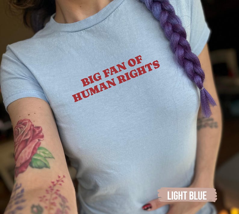 Op de afbeelding: Lichtblauw T-shirt met de rode tekst "BIG FAN OF HUMAN RIGHTS". Het shirt heeft een ronde hals. De afbeelding toont een close-up van het shirt.