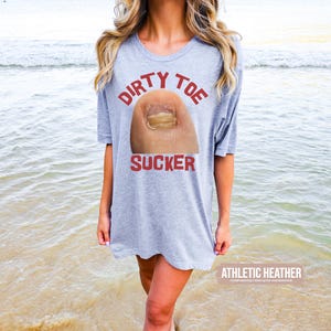 Pu&ograve; includere: Una maglietta grigio chiaro con le parole "DIRTY TOE SUCKER" in rosso sopra una grafica di un'unghia del piede. Le parole "ATHLETIC HEATHER" sono stampate in bianco in fondo alla maglietta. La persona &egrave; su una spiaggia.