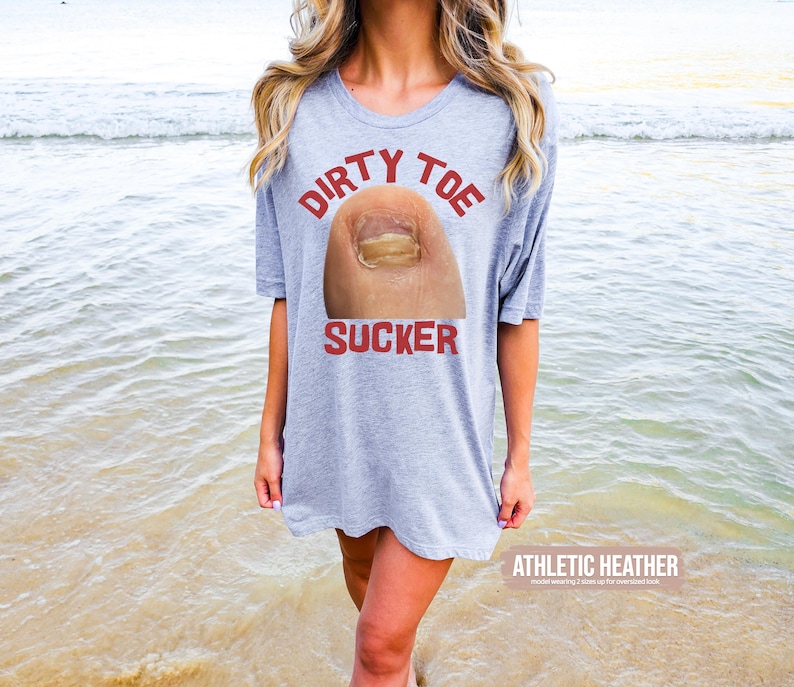 Pu&ograve; includere: Una maglietta grigio chiaro con le parole "DIRTY TOE SUCKER" in rosso sopra una grafica di un'unghia del piede. Le parole "ATHLETIC HEATHER" sono stampate in bianco in fondo alla maglietta. La persona &egrave; su una spiaggia.