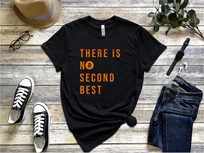 May include: Black t-shirt with the text "THERE IS NO SECOND BEST" in orange lettering. A Bitcoin symbol is incorporated into the text. The shirt is laid flat on a wooden surface with a pair of black sneakers, a straw hat, a watch, and a pair of jeans.