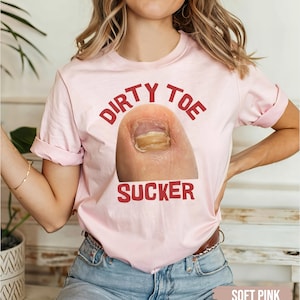Pu&ograve; includere: T-shirt rosa chiaro con la scritta rossa "DIRTY TOE SUCKER" e una foto di un dito del piede con un'infezione fungina. La maglietta &egrave; di colore rosa tenue.