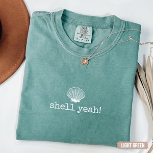 May include: Light green t-shirt with the text "shell yeah!" and a seashell graphic. A gold necklace with a pendant is draped across the shirt. The shirt is folded and placed on a white surface.