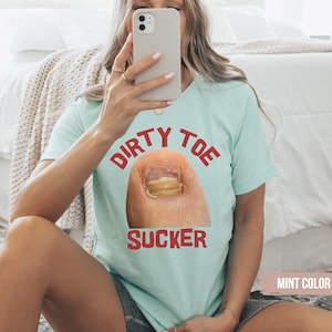 Pu&ograve; includere: Una maglietta color menta con le parole "DIRTY TOE SUCKER" in lettere rosse sopra e sotto un'immagine in primo piano di un'unghia del piede. La maglietta &egrave; tenuta da una persona.