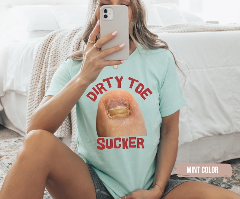 Pu&ograve; includere: Una maglietta color menta con le parole "DIRTY TOE SUCKER" in lettere rosse sopra e sotto un'immagine in primo piano di un'unghia del piede. La maglietta &egrave; tenuta da una persona.