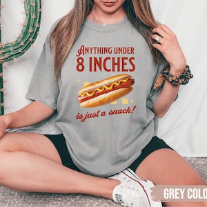 May include: A gray t-shirt with the text "Anything under 8 inches is just a snack!" and a graphic of a hot dog with mustard. The shirt is a casual style.