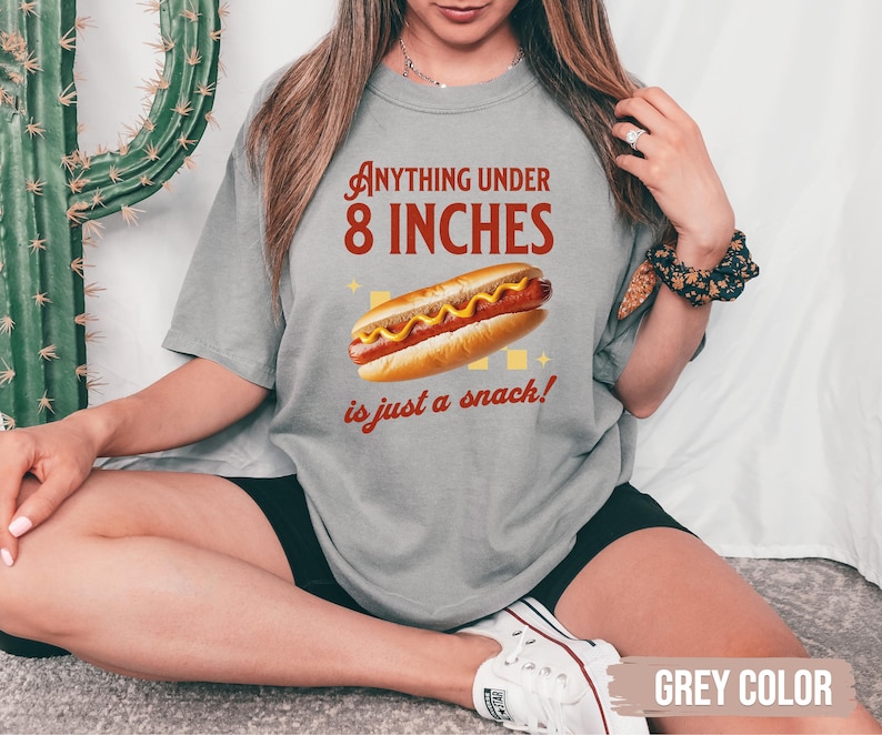 May include: A gray t-shirt with the text "Anything under 8 inches is just a snack!" and a graphic of a hot dog with mustard. The shirt is a casual style.