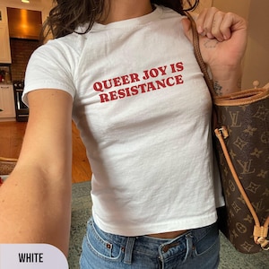 May include: White t-shirt with the red text "QUEER JOY IS RESISTANCE". The shirt is paired with blue jeans. A brown handbag with a monogram pattern is visible.