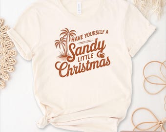 May include: A cream-coloured t-shirt with rolled-up sleeves. The shirt displays the text "Have Yourself a Sandy Little Christmas" in brown, accompanied by palm trees and a beach ball graphic. The words "Natural Color" are at the bottom.